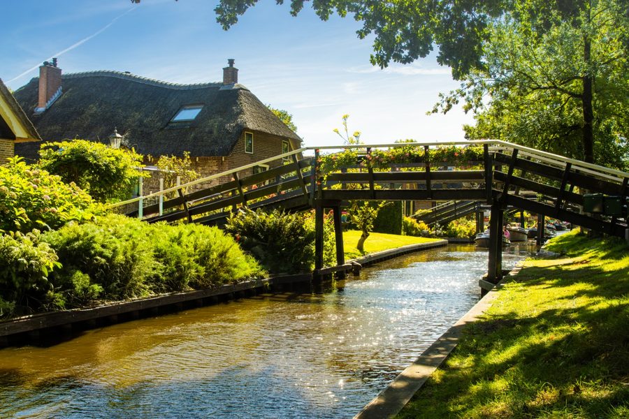 Houten bruggetje over een gracht met een rietgedekt huis en groene tuinen in Giethoorn