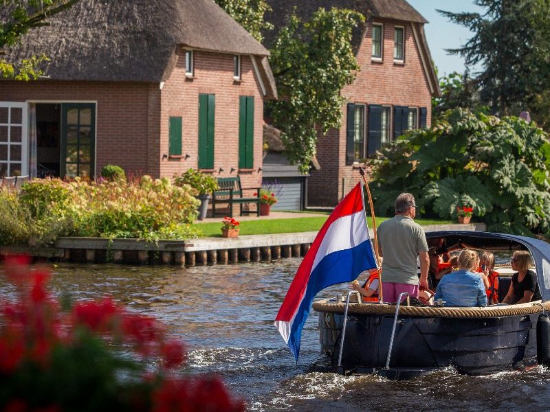 Boot mit Skipper und niederländischer Flagge fährt an reetgedeckten Häusern in Giethoorn vorbei