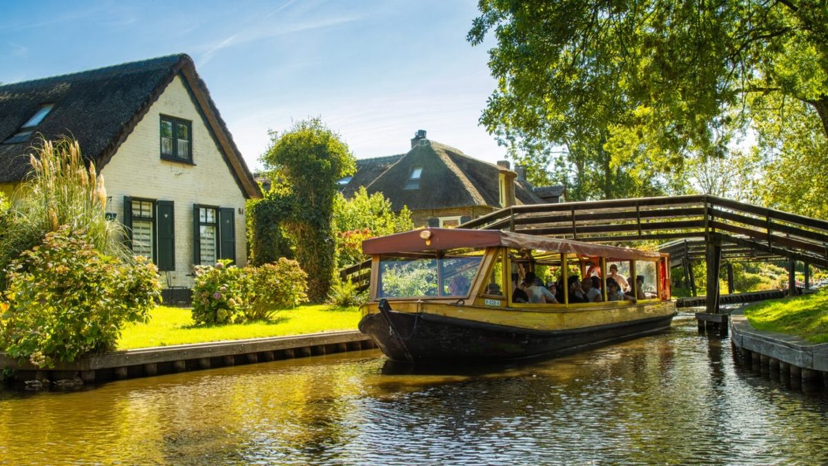 Rundfahrtboot mit geöffneten Fenstern fährt durch eine Gracht in Giethoorn an einem reetgedeckten Bauernhof vorbei