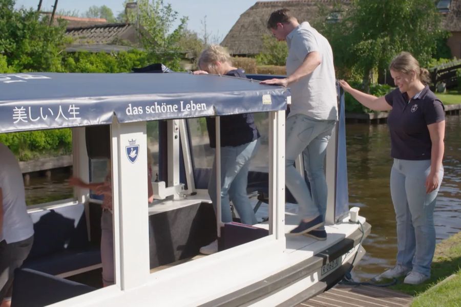 Gäste steigen in ein überdachtes Rundfahrtboot von Boatrental Giethoorn ein, unterstützt von einer Mitarbeiterin