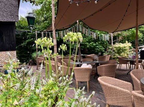 Terrasse von De Landije mit Korbstühlen unter einem Tuch zwischen blühenden Pflanzen