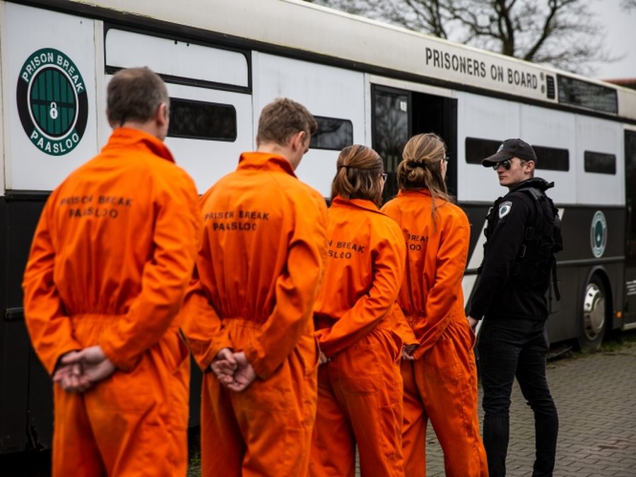 Players in orange prison overalls with a guard by the bus of Prison Break Paasloo