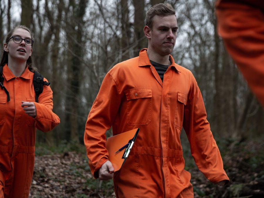 Two players in orange overalls walk through the forest with a clipboard during Prison Break Paasloo