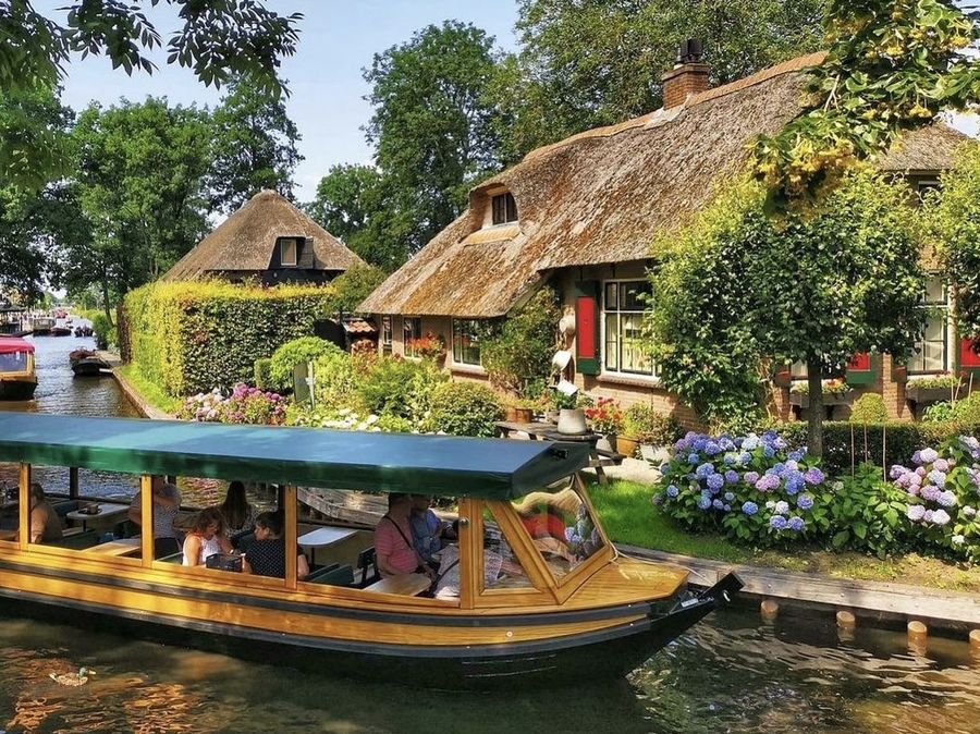 Rundfahrtboot mit Fahrgästen fährt an Höfen mit Reetdächern und Hortensien in Giethoorn vorbei
