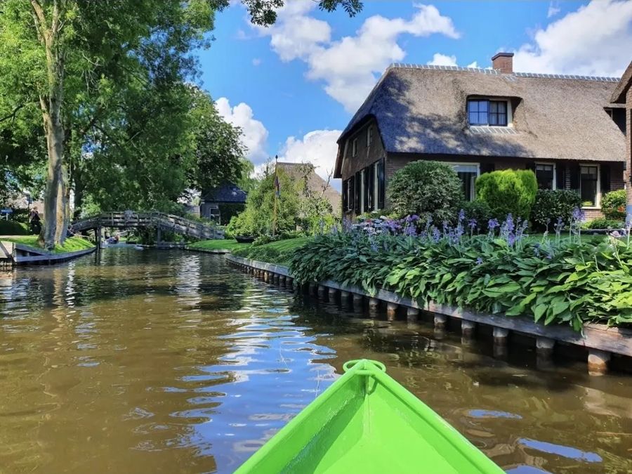 Blick vom Bug eines grünen Mietbootes auf eine Gracht mit einem Hof mit Reetdach