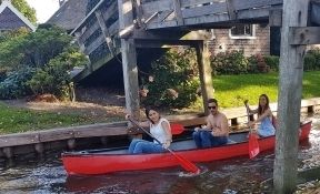 Drei Personen fahren in einem roten Kanu unter einer Holzbrücke hindurch in Giethoorn